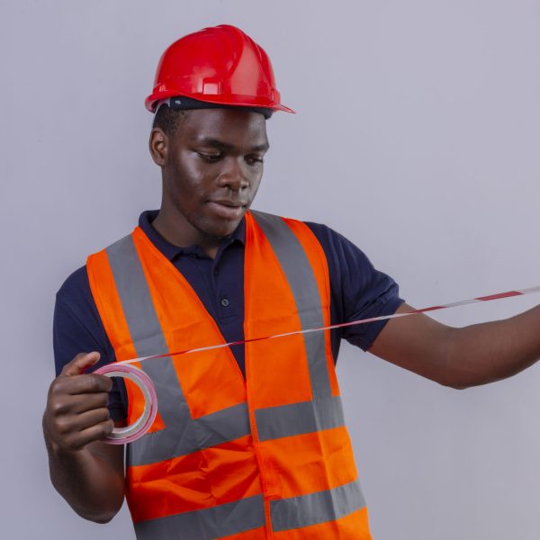 young african american builder man wearing construction vest and safety helmet looking at using measuring tape looking at it standing over white background